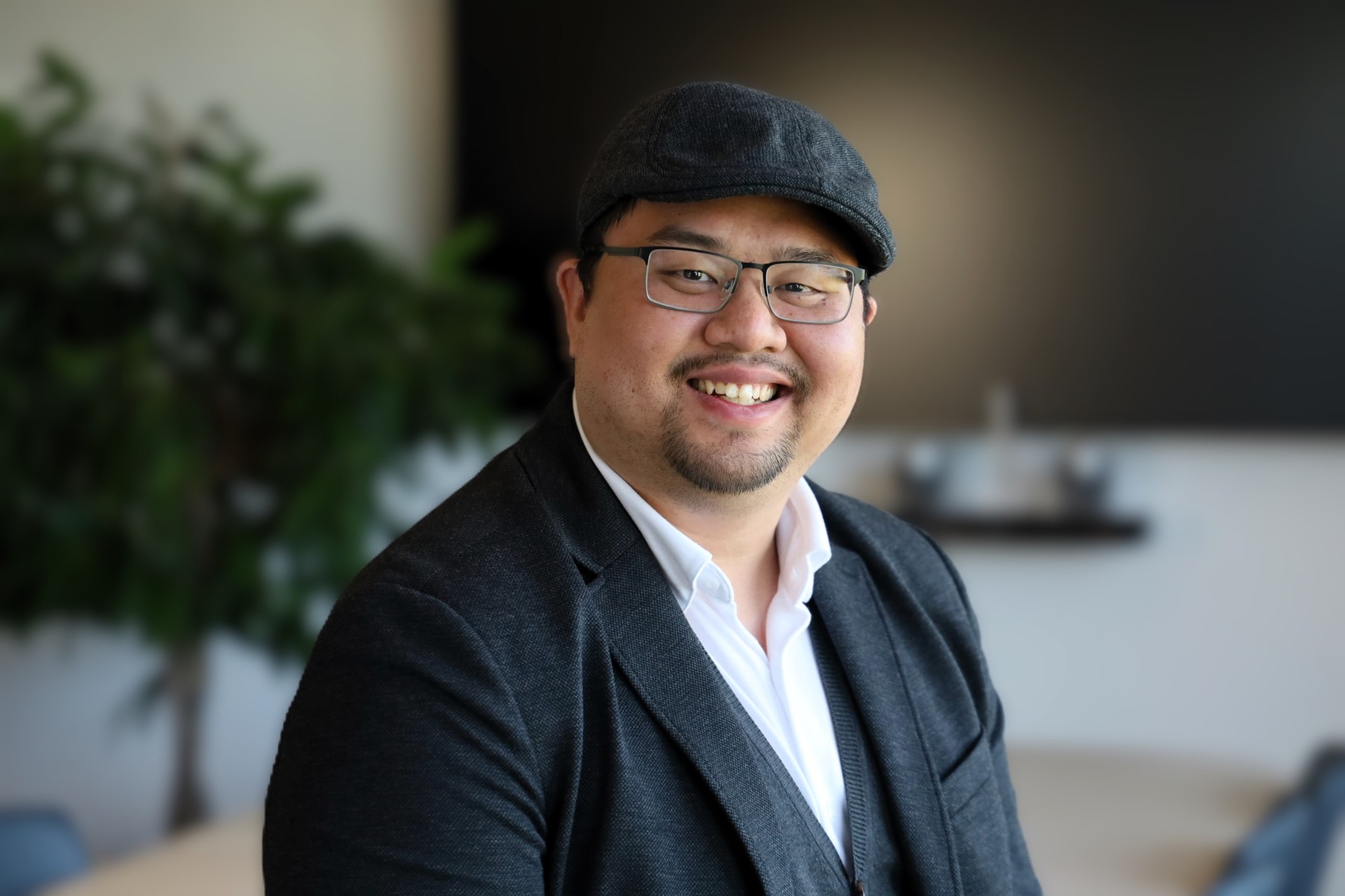 Portrait of Joakim Cao, smiling, wearing a black cap, glasses and a dark blazer.
